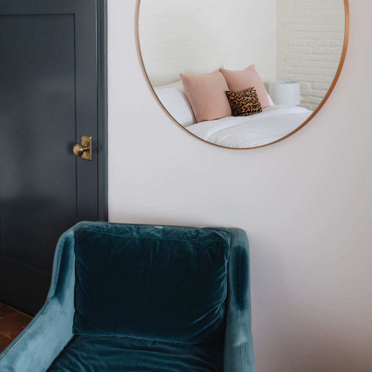 A round mirror is mounted on a white brick wall next to a dark door, while a teal velvet armchair is positioned nearby.