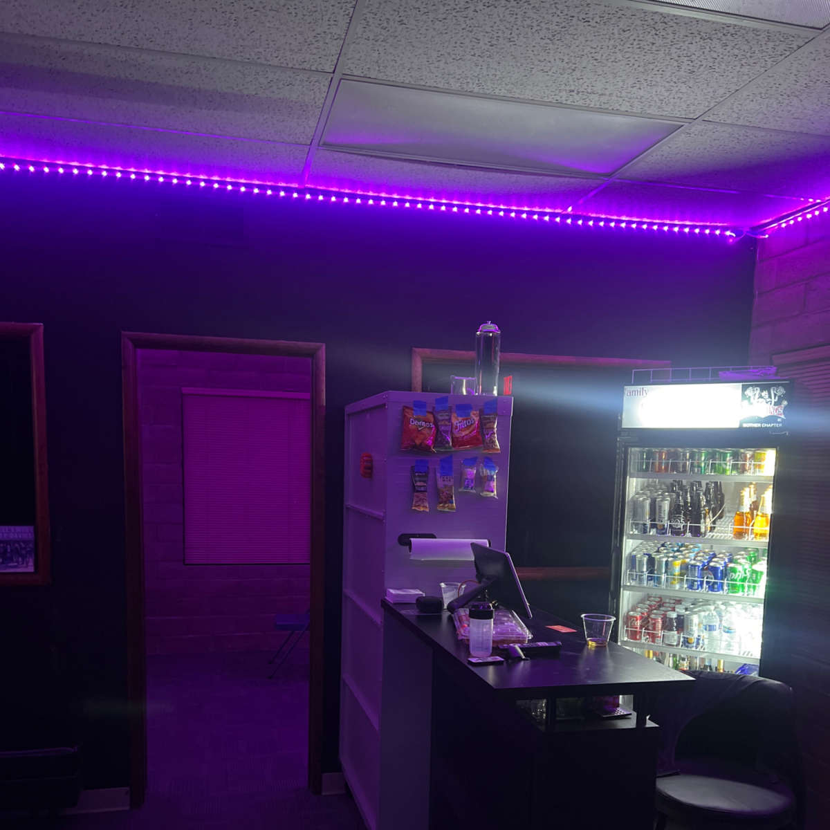 A dimly lit room with purple LED lighting, featuring a bar area with a cash register and a refrigerator stocked with beverages.