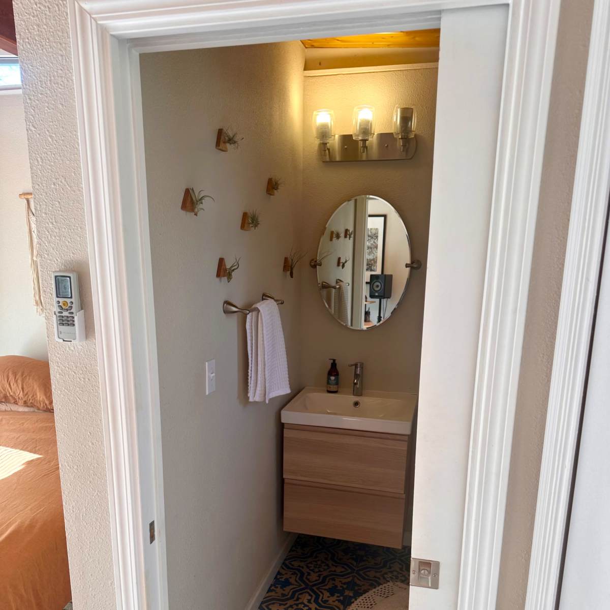 A bathroom entrance featuring a wooden vanity with a round mirror, a wall-mounted towel rack, and decorative plants on the wall, with patterned flooring visible.