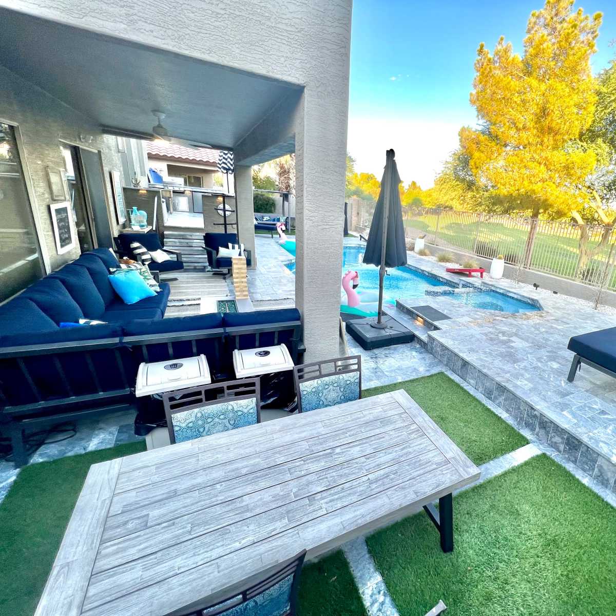 A backyard patio with a pool, a seating area with a large sofa, a dining table, and a sun lounger surrounded by greenery.
