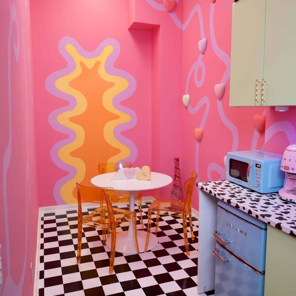 A colorful kitchen with pink walls, a checkered black-and-white floor, and a round table surrounded by transparent chairs.