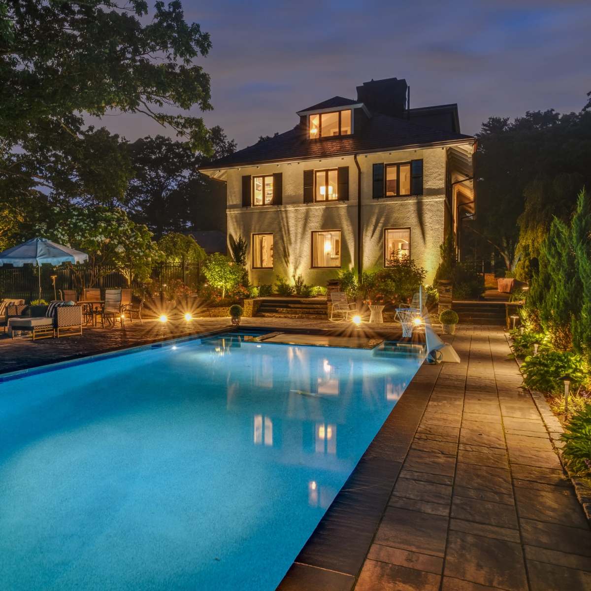 A well-lit backyard features a swimming pool beside a two-story house with illuminated windows and landscaped greenery.