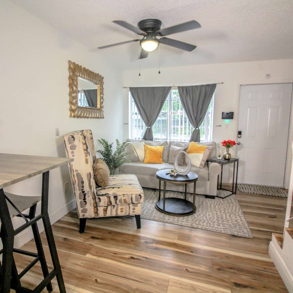 A small living area with a couch, chairs, a table, and decorative elements, featuring large windows and a staircase.