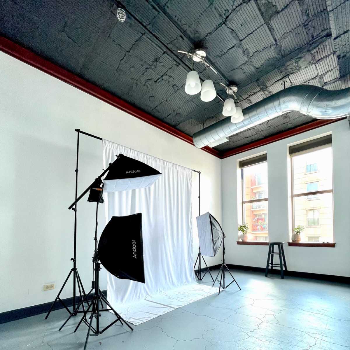 A photography studio setup with a white backdrop, lighting equipment, and a stool in a room with large windows.