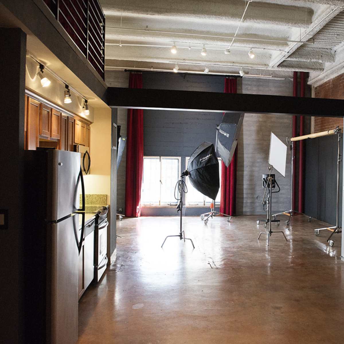 An industrial-style interior with a kitchen on the left and photography equipment set up on the right, including softboxes and a backdrop.