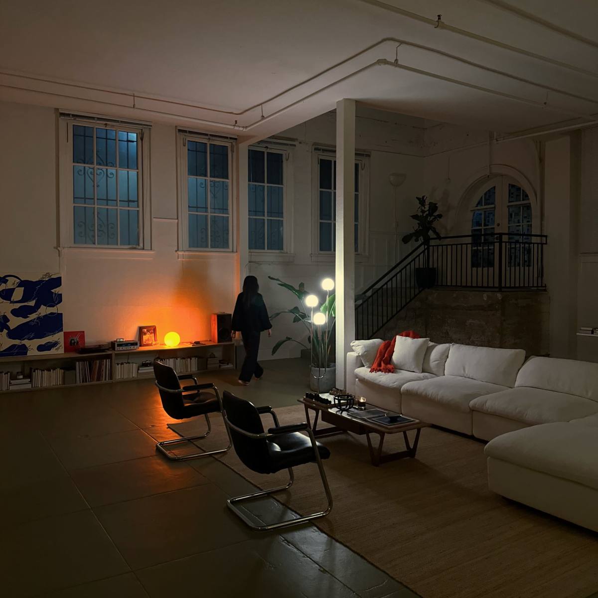 A person stands near a modern white sofa in a spacious, dimly lit living room with large windows and minimalist decor.