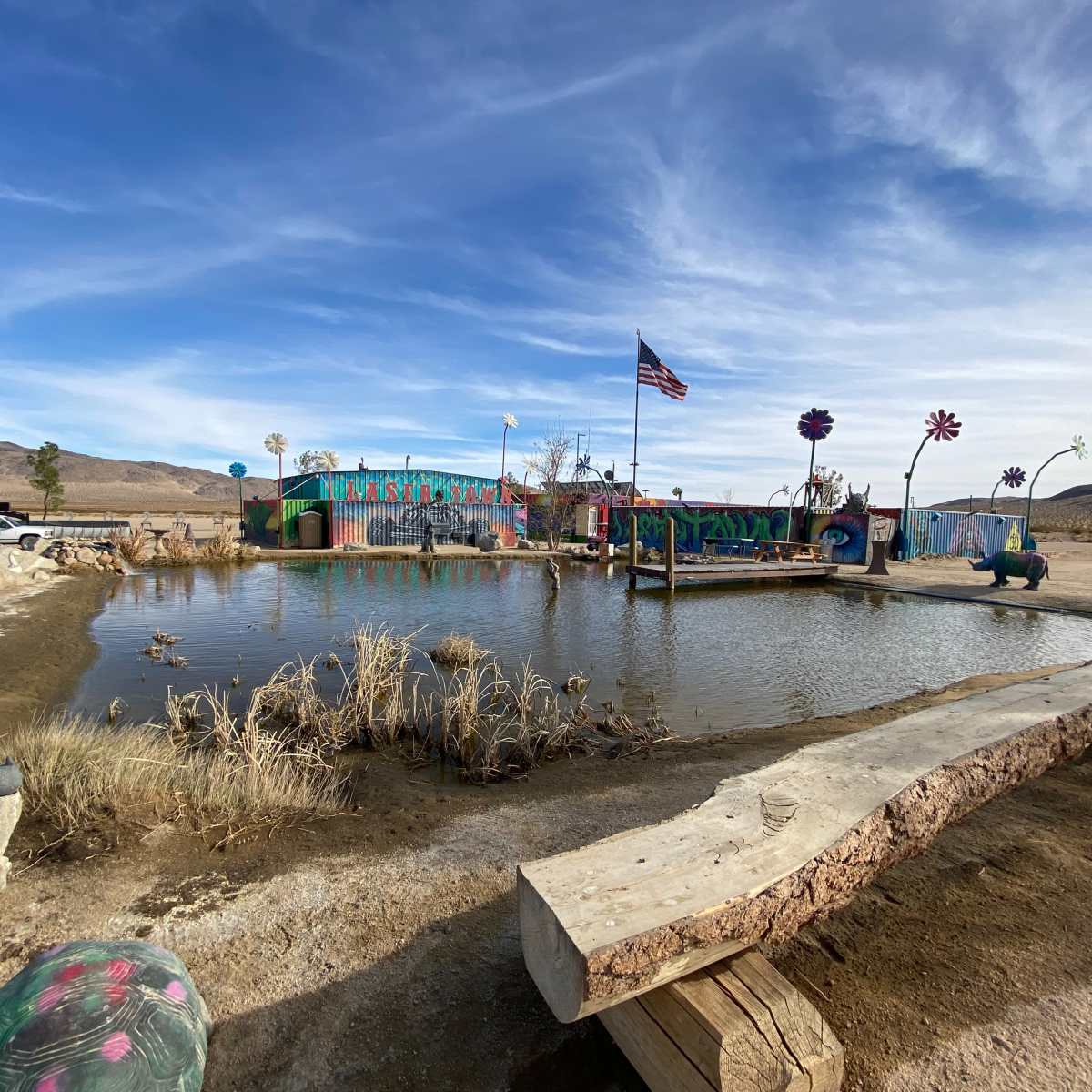 A colorful building with artistic murals is situated beside a pond in a desert landscape.