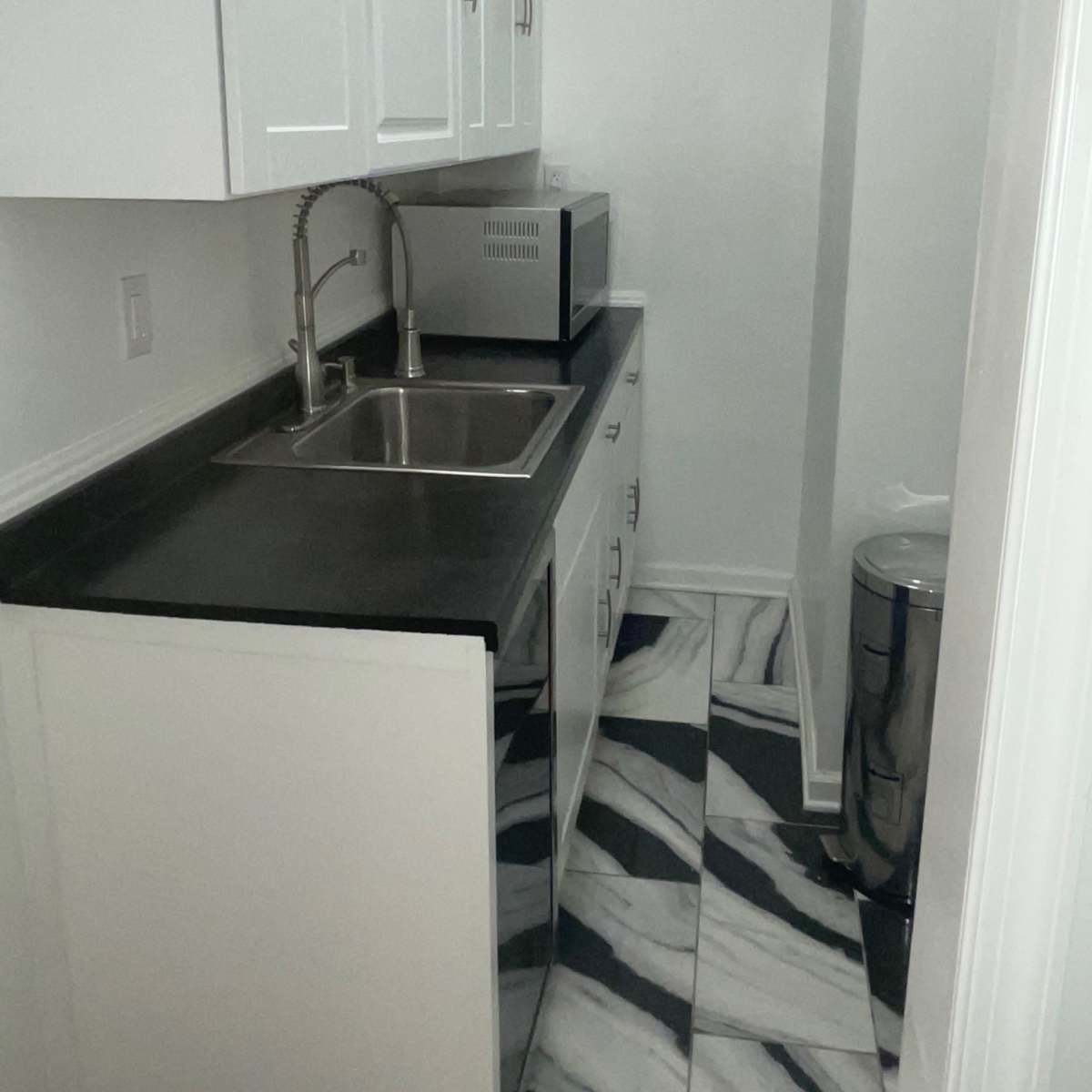 A narrow kitchen space with white cabinets, a stainless steel sink, a countertop, a microwave, and a trash can against a marbled floor.
