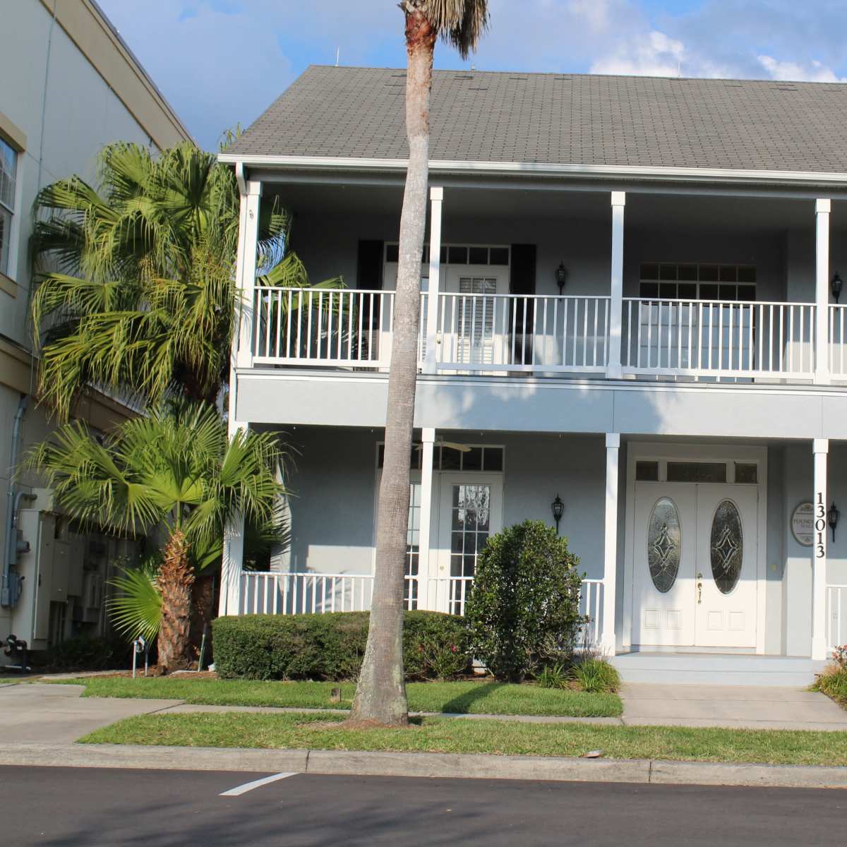 A two-story blue house with white railings and double front doors, flanked by landscaping and palm trees.