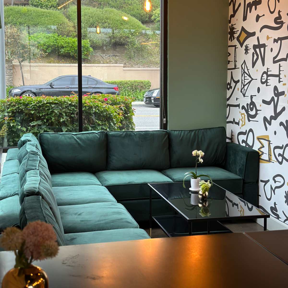 A large green sectional couch is positioned in a well-lit corner of a modern space, next to a glass coffee table and a large window displaying greenery and a street view.
