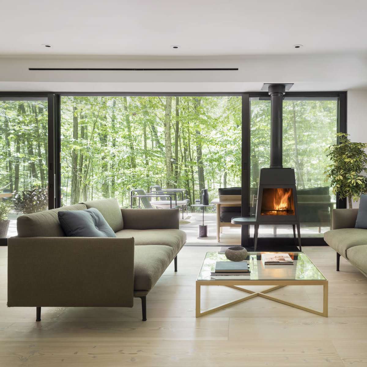 A modern living room with two green sofas facing a glass coffee table and a wood-burning stove, surrounded by large windows that overlook a forested area.