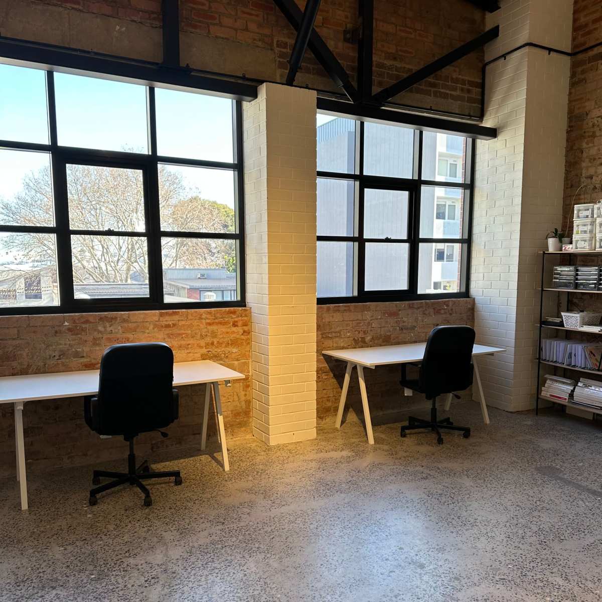 A spacious room with two white desks and black chairs positioned next to large windows, featuring brick walls and a polished concrete floor.