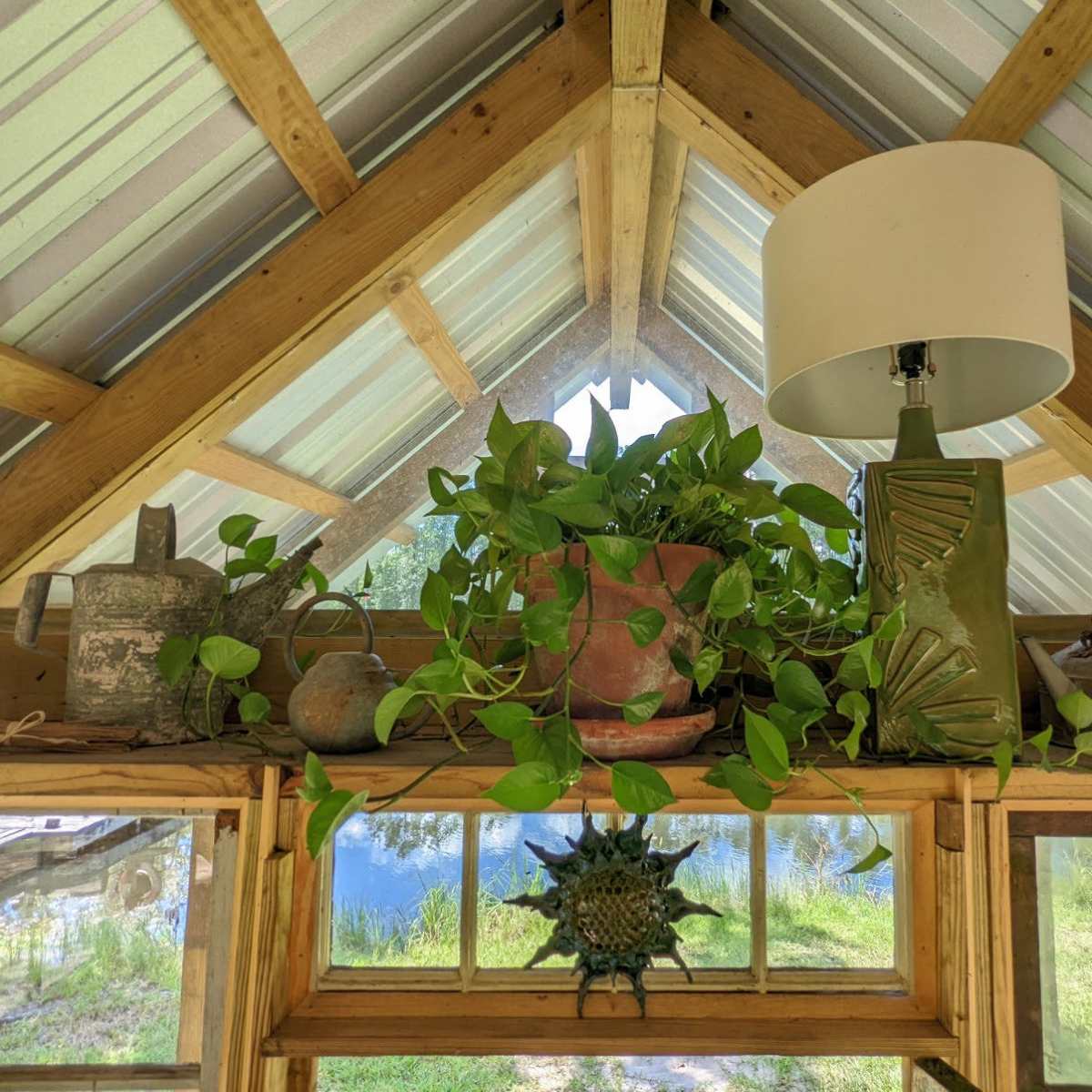 A shelf in a greenhouse holds potted plants, decorative pottery, and a lamp under a slanted roof.