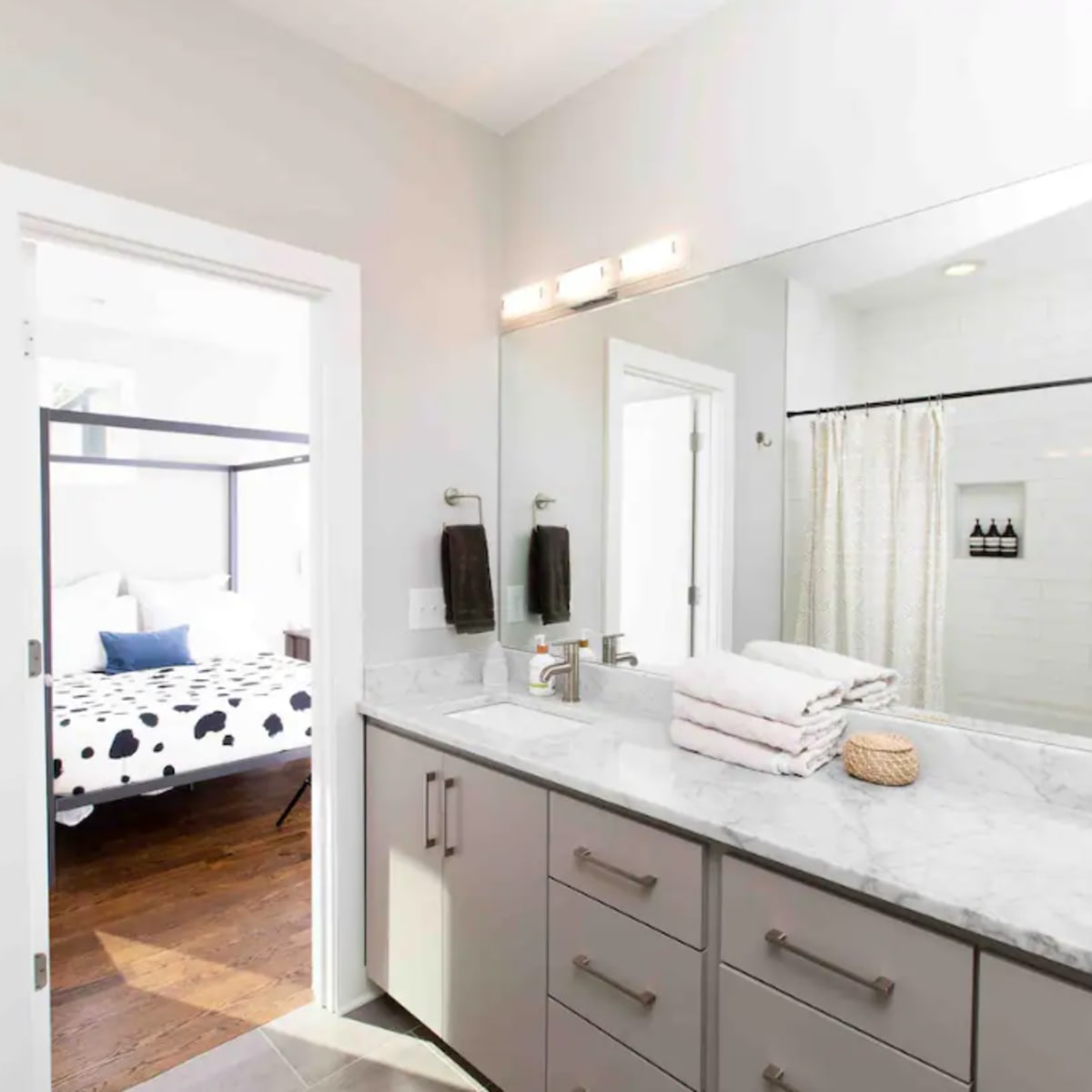 A modern bathroom features a double vanity with a marble countertop and a doorway leading to a bedroom with a four-poster bed.