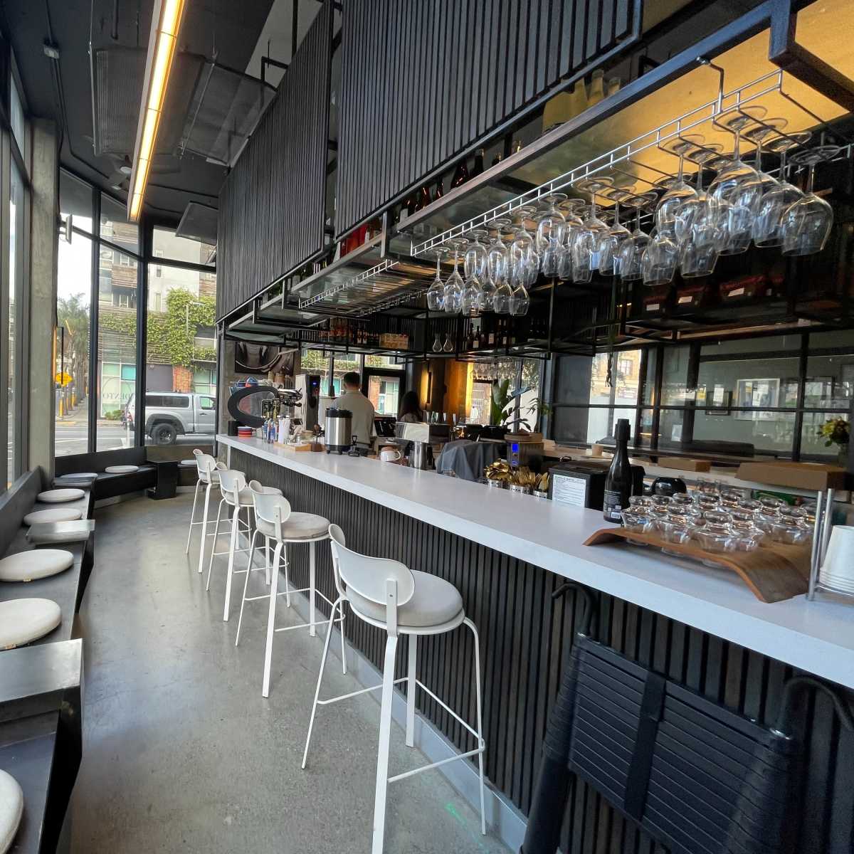 A modern restaurant interior with a bar area featuring high stools, a countertop, and large windows overlooking the street.
