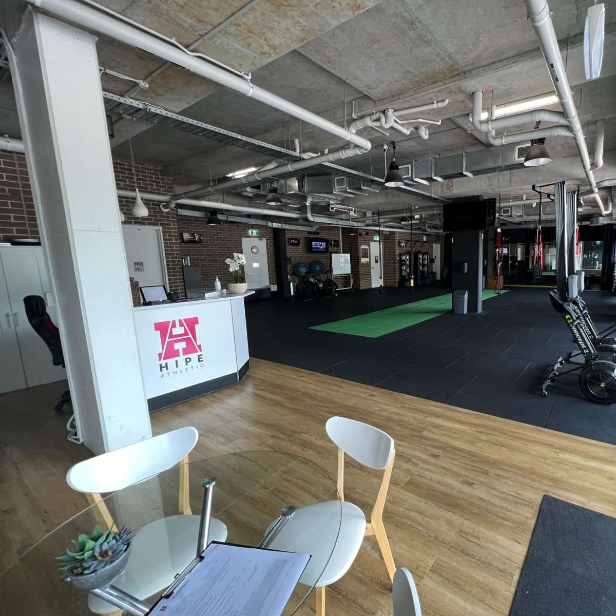 A modern fitness facility featuring an open gym area with exercise equipment, a reception desk, and wooden flooring.