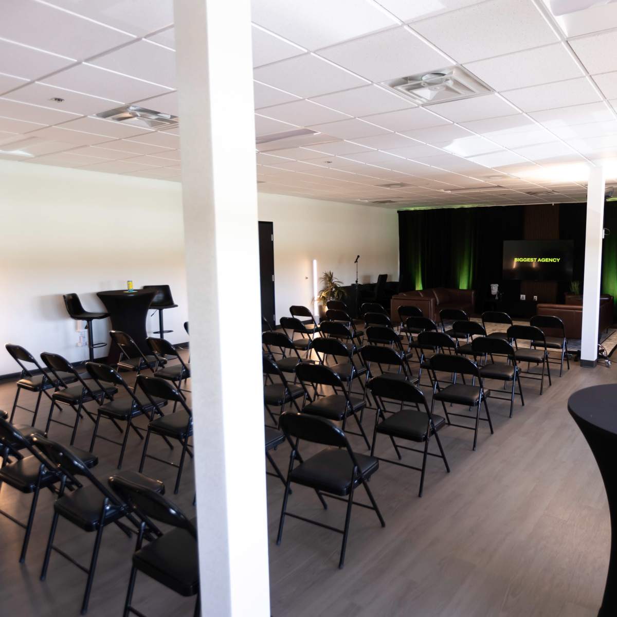 A spacious room with black chairs arranged in rows facing a stage, accompanied by high-top tables and green lighting.