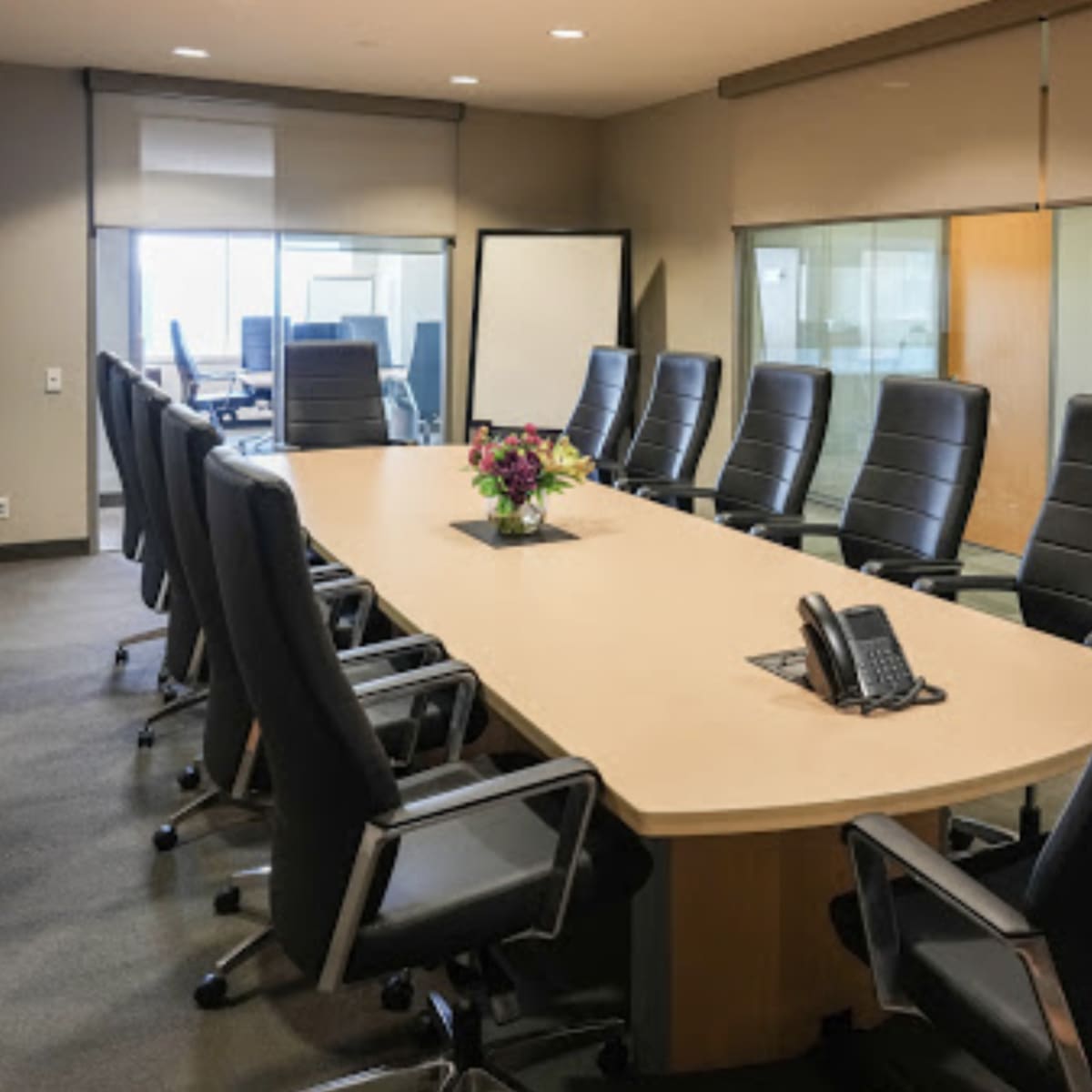 A large, modern conference room features a long, oval table surrounded by black leather chairs, with a floral centerpiece and a phone placed at the center.