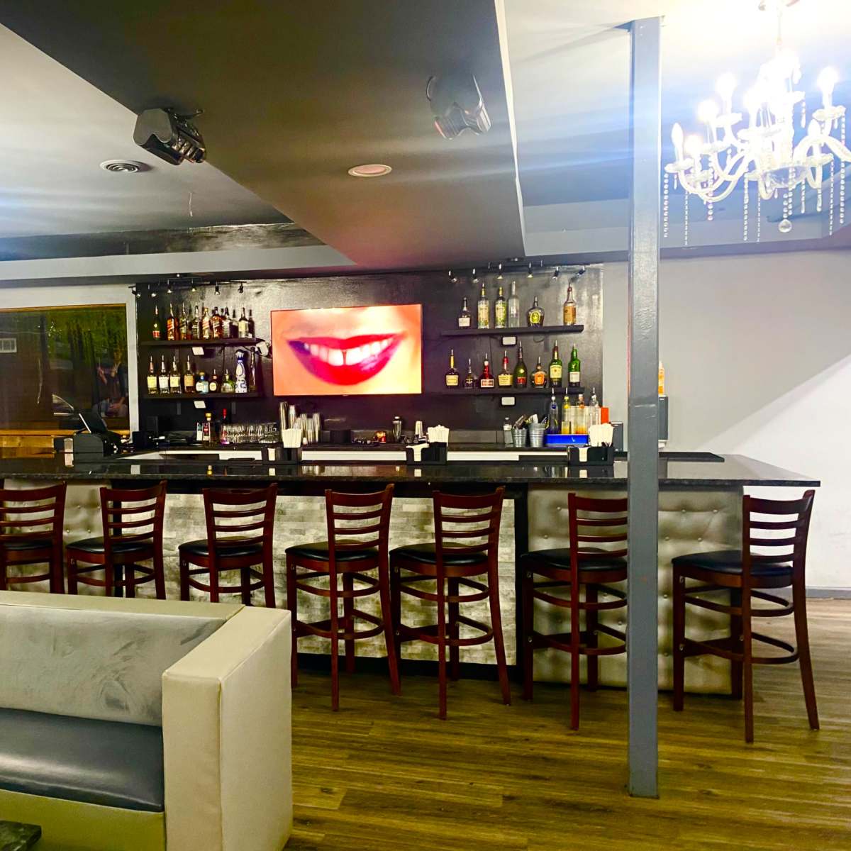 A bar area features a long counter with high chairs, illuminated by a large image of a smiling mouth on the wall behind the shelves of liquor.