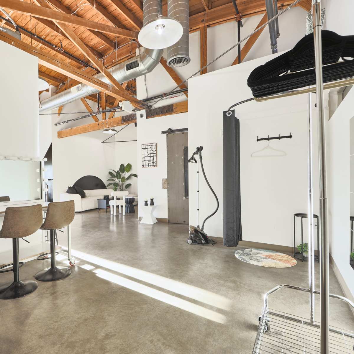 A modern studio space with a concrete floor, exposed wooden beams, a seating area with mirrors, and various equipment along the walls.