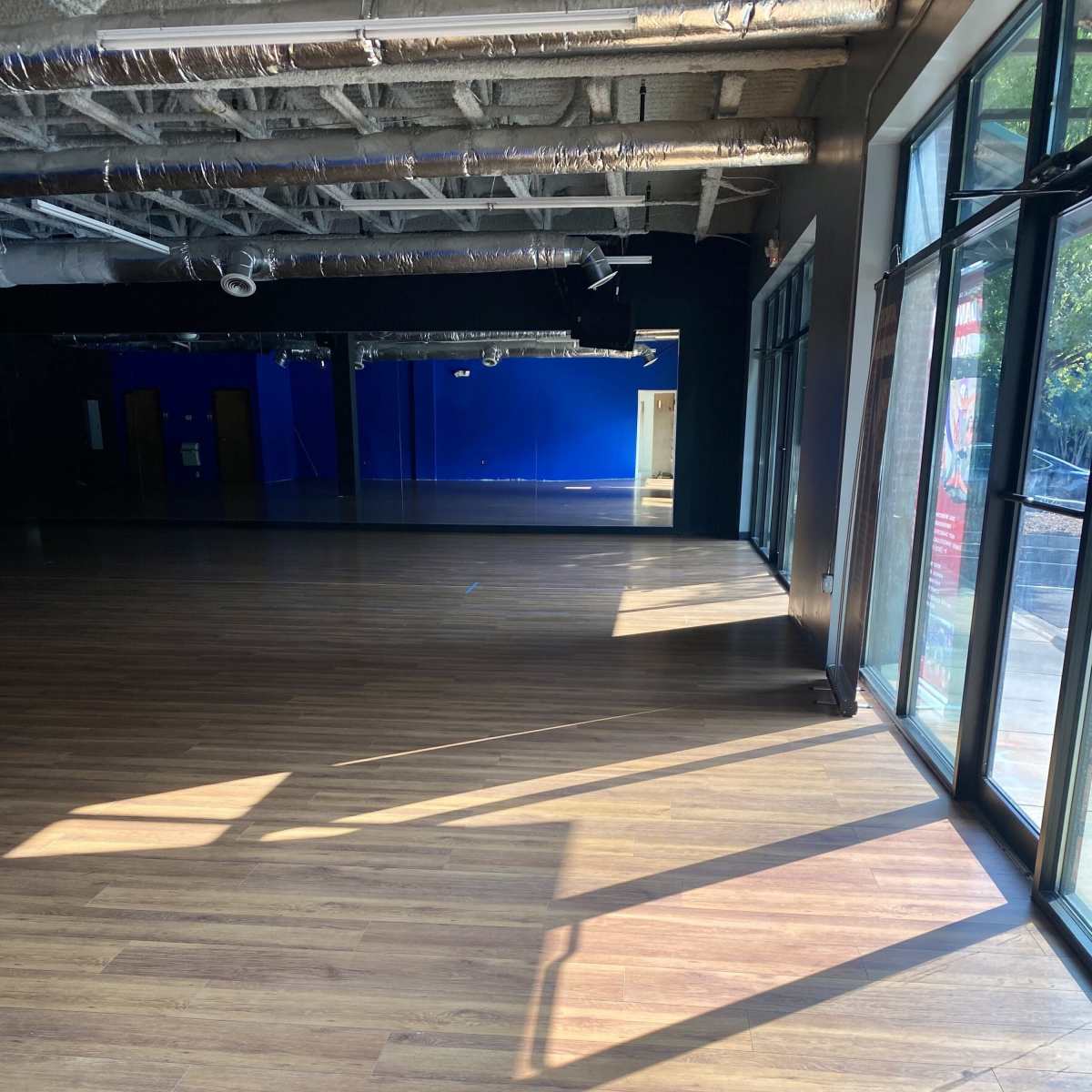 A spacious, empty room with wooden flooring and large windows allowing sunlight to create shadows on the floor.