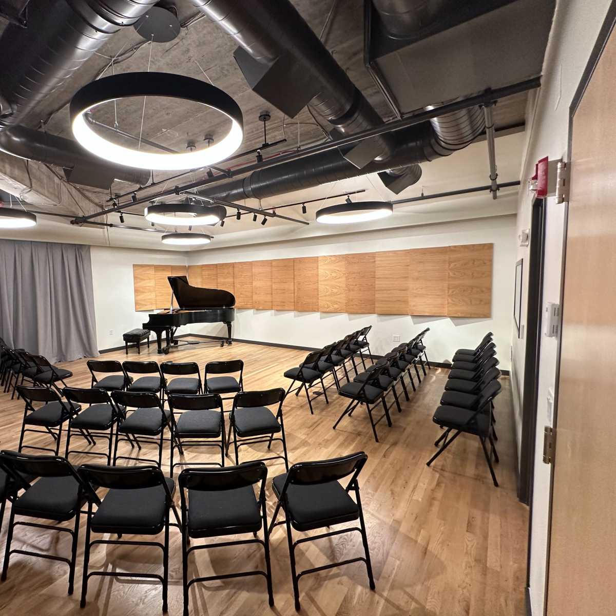 A small performance space features a grand piano, surrounded by neatly arranged black chairs and wooden paneling on the wall.