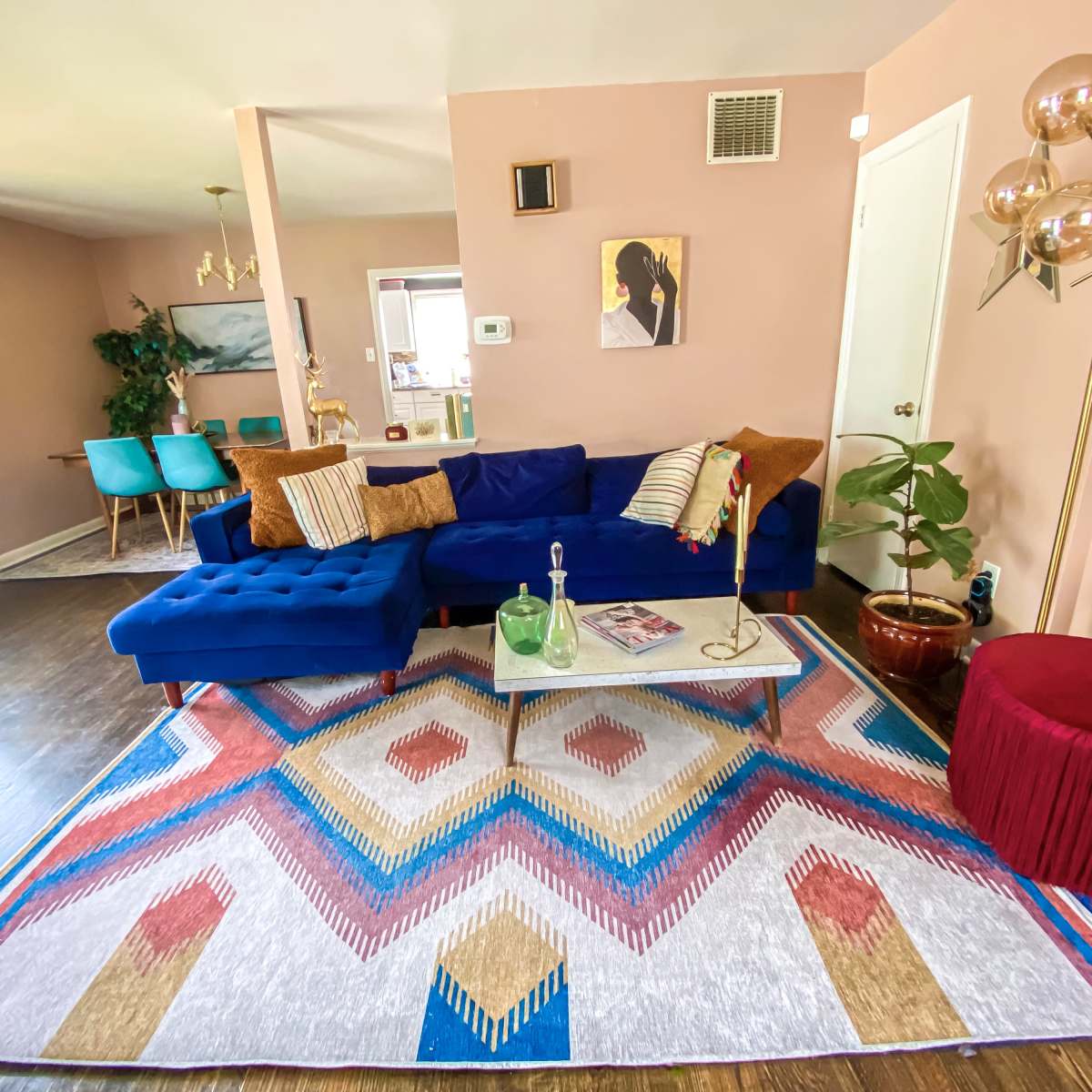 A bright living room featuring a blue sectional sofa, a geometric area rug, a glass coffee table, and a mix of decorative plants and furniture.