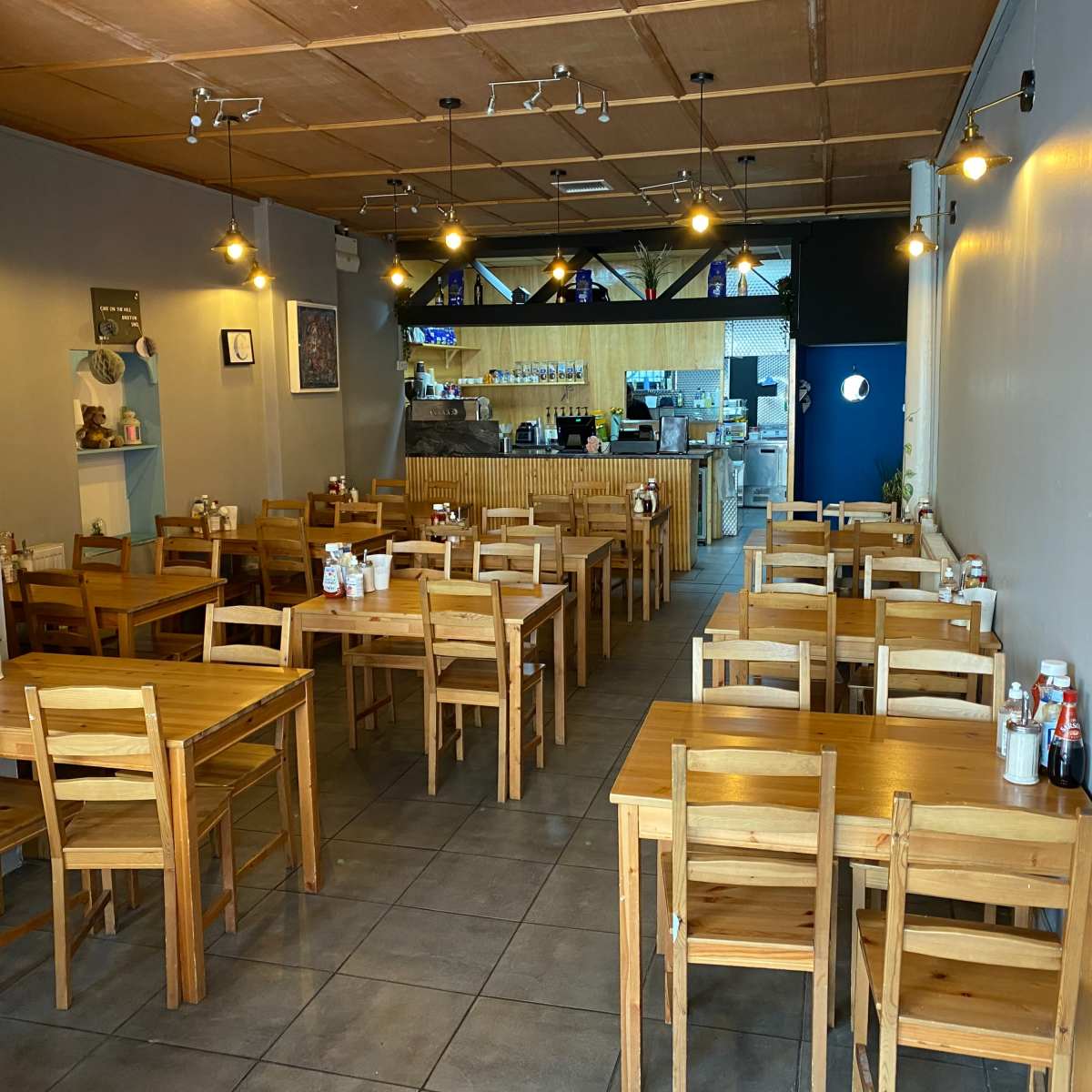 A spacious, lightly furnished restaurant with wooden tables and chairs, and a counter visible at the back.