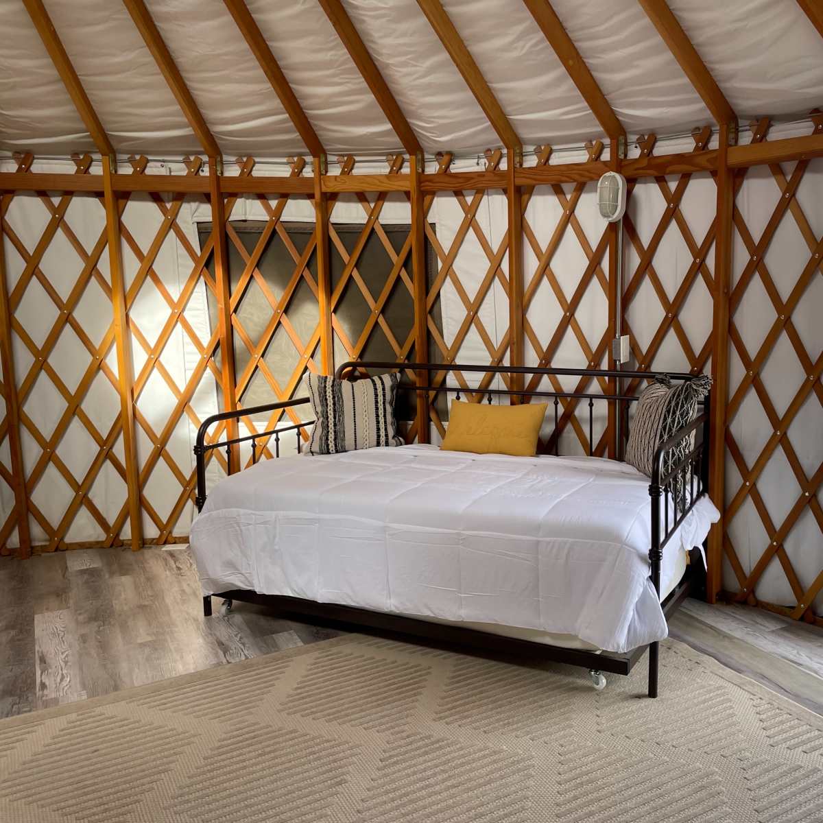 The interior of a yurt features a daybed with a white comforter, flanked by patterned pillows, and large windows showing a view of the landscape outside.