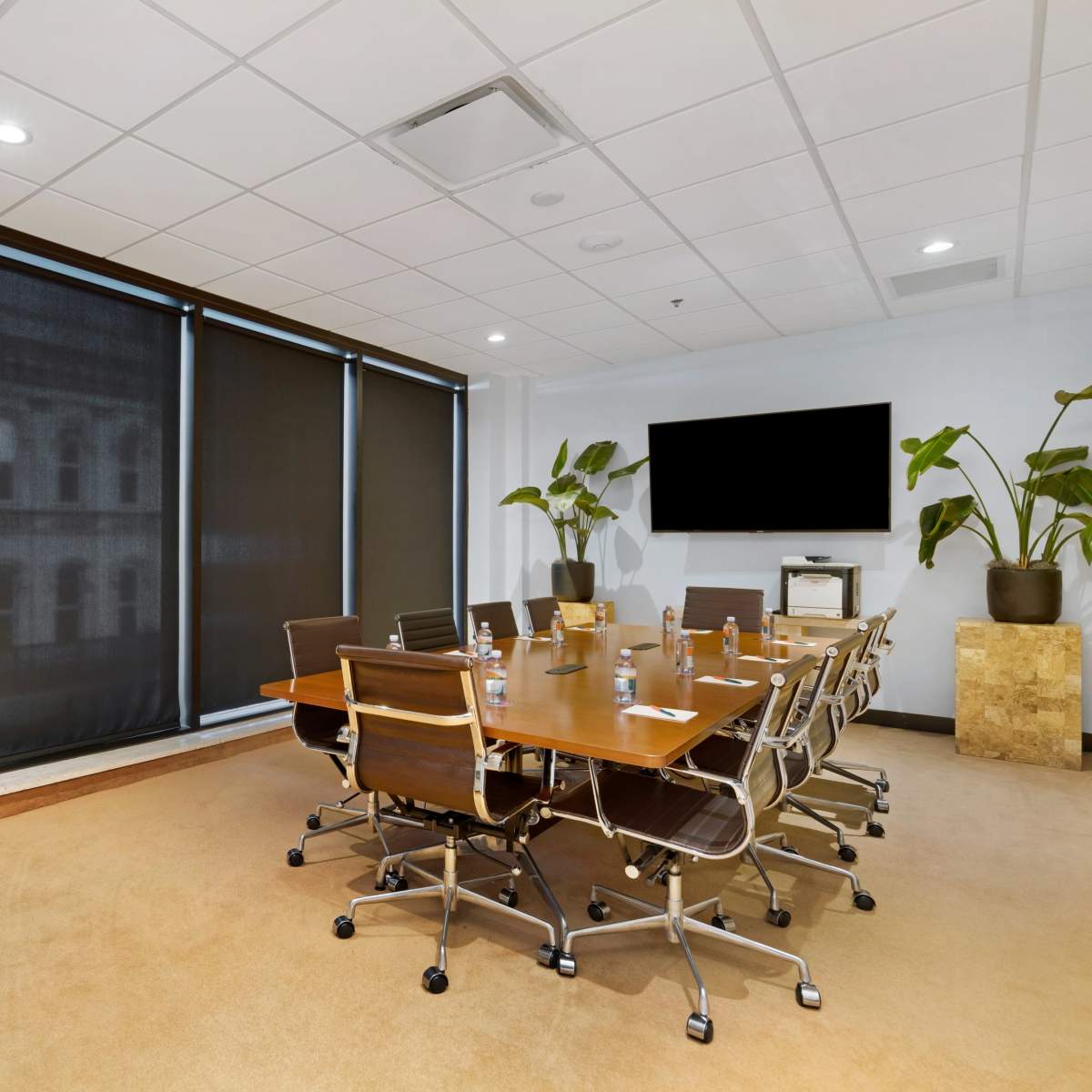 A modern conference room features a large table surrounded by ergonomic chairs, with dark window shades and a wall-mounted screen.