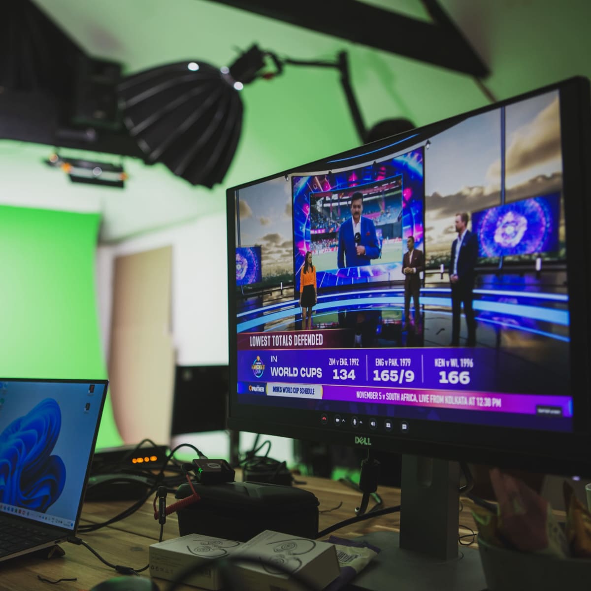 A computer monitor displaying a sports news broadcast is positioned on a desk with various equipment, while a presenter stands in front of a green screen in the background.