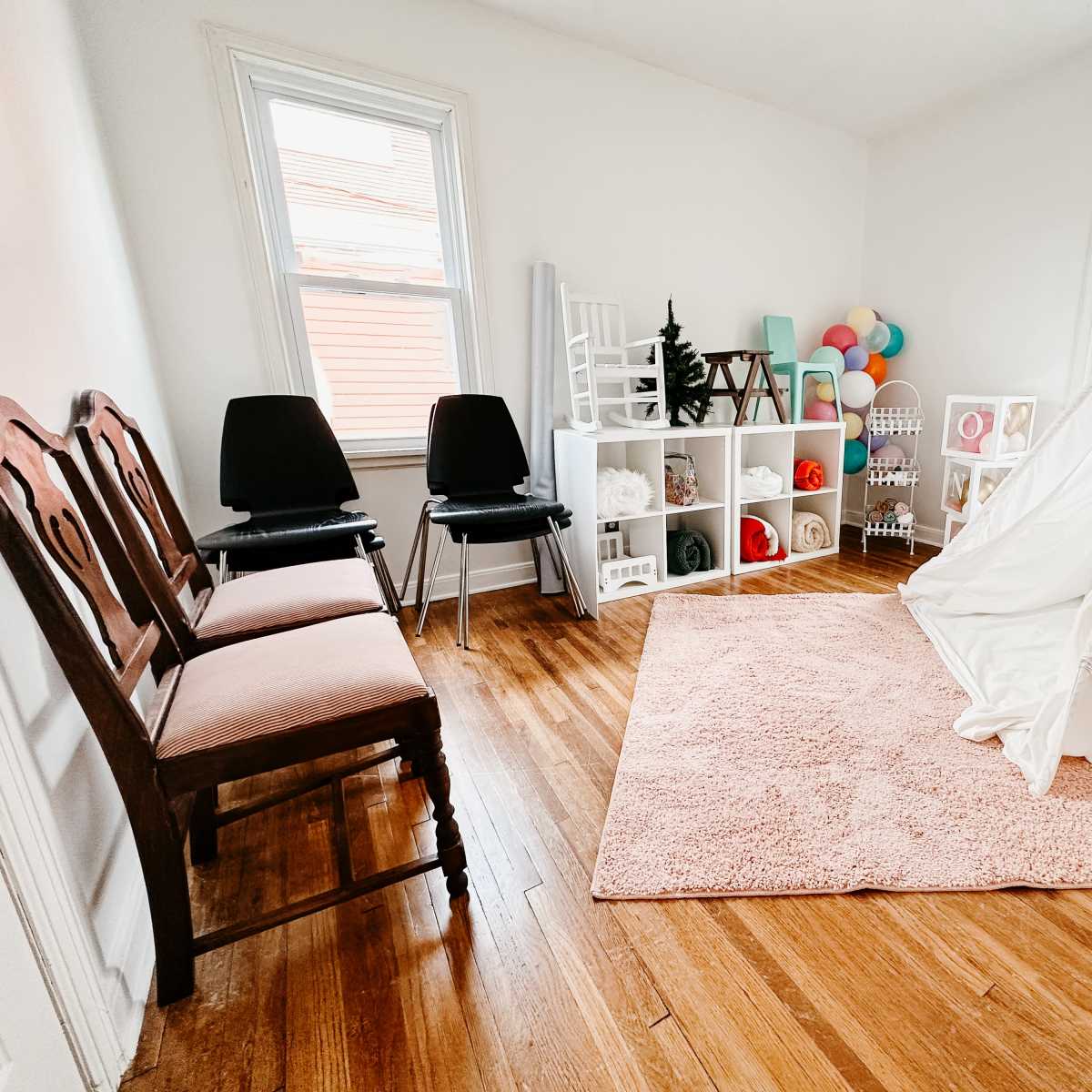 A bright room with two chairs, a white play tent, a soft pink rug, and storage shelves filled with colorful toys and decorations.