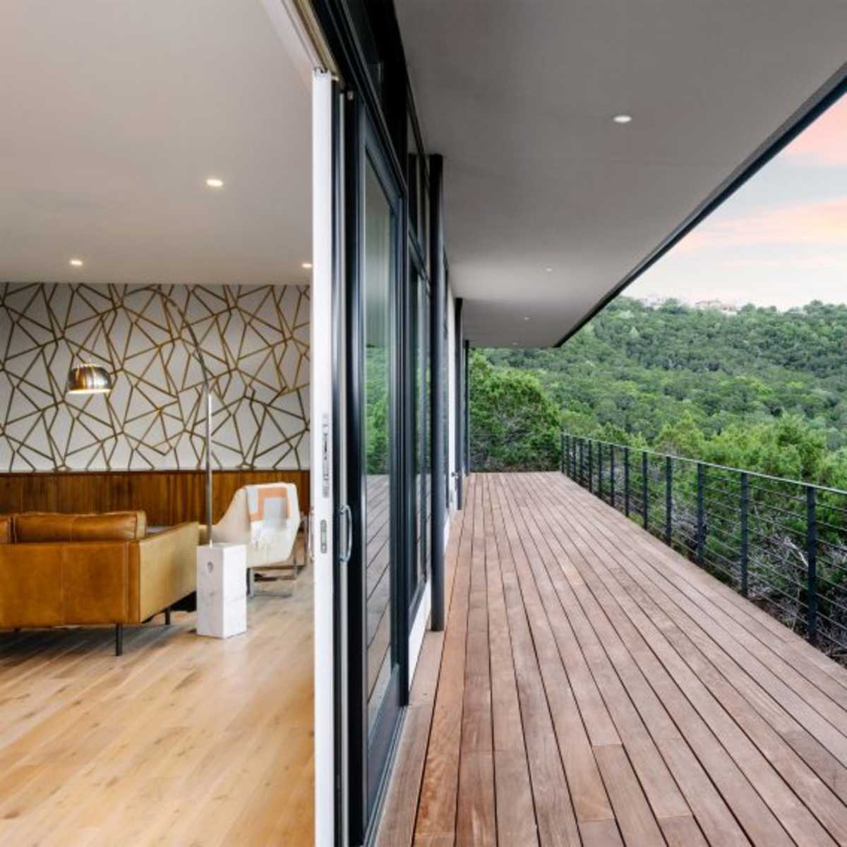 A wooden deck extending from a modern interior space, overlooking a green hillside under a colorful sky.