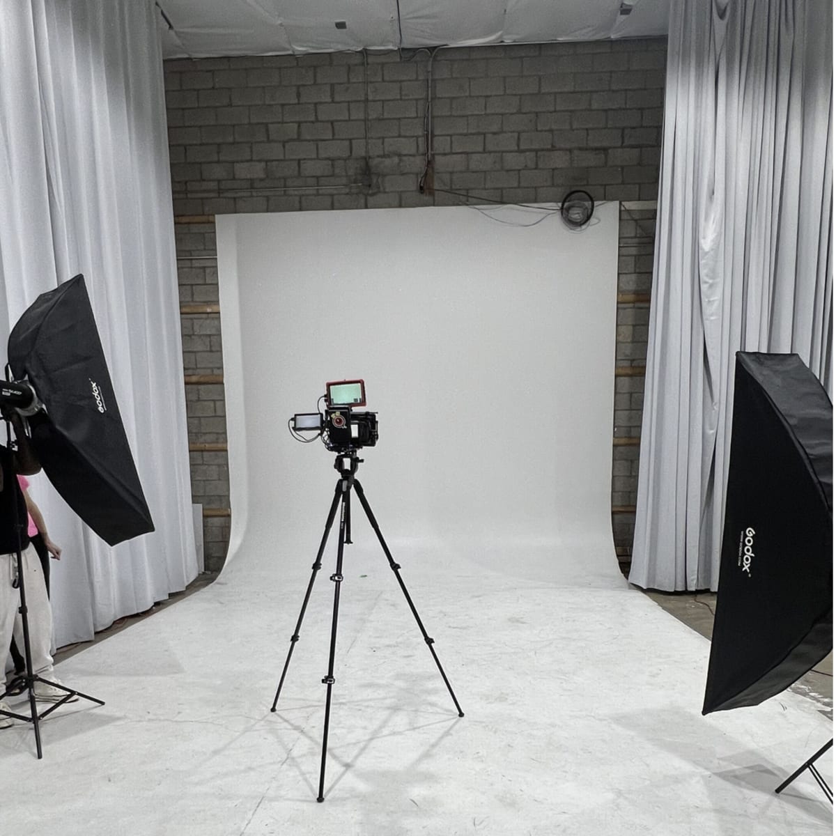 A photography studio setup with a white backdrop, a camera on a tripod, and two softbox lights positioned on either side.