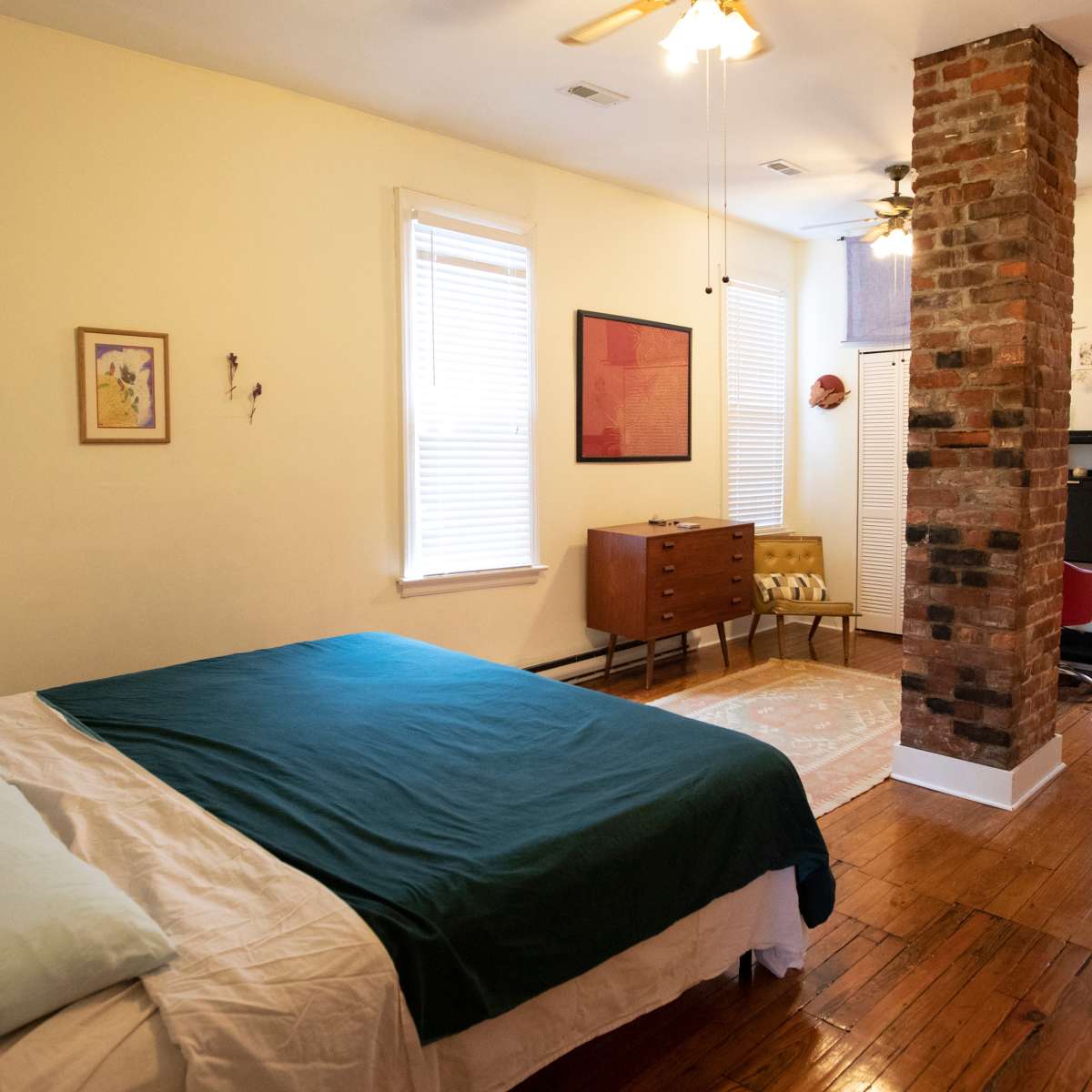 A bedroom features a bed with a green blanket, a wooden dresser, and exposed brick accents.