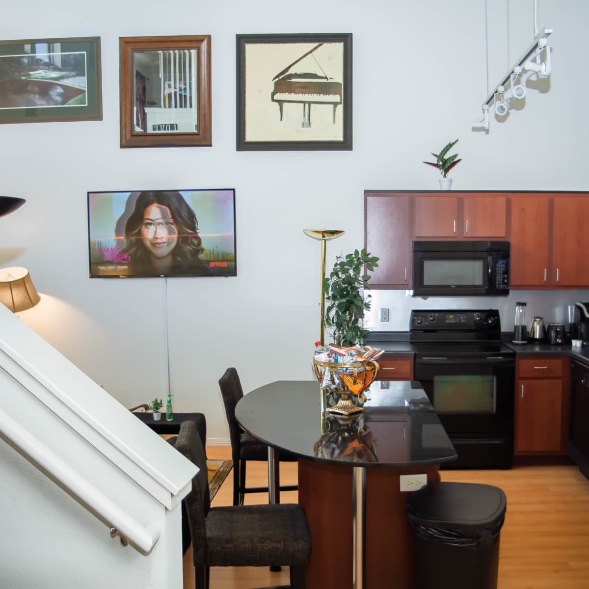 A modern kitchen with dark wood cabinetry and appliances, a round table with chairs, and a living area featuring a television displaying a woman.