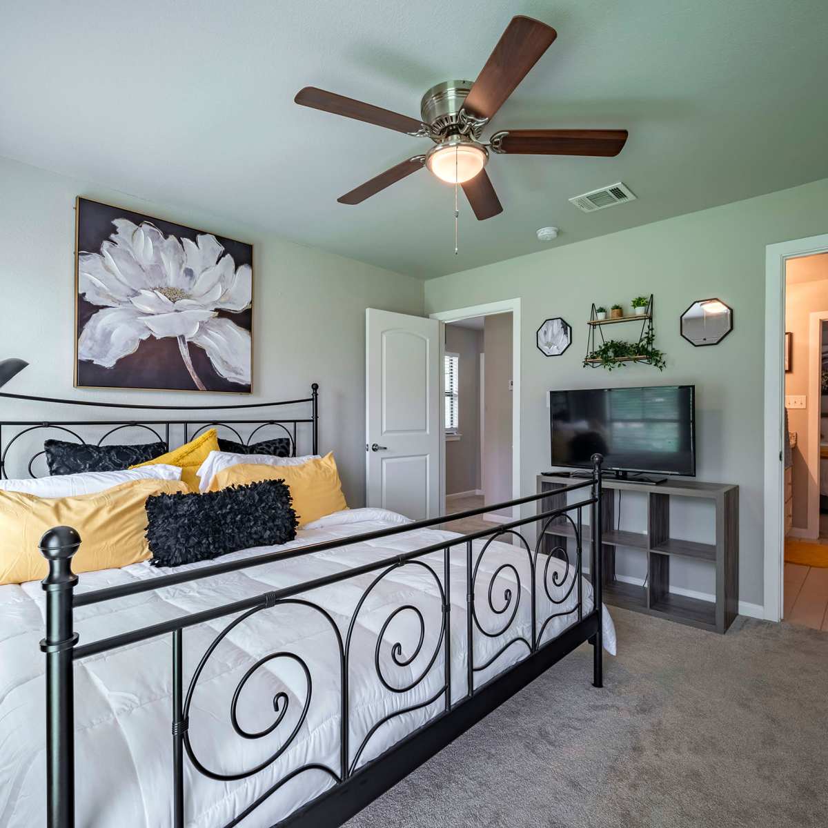 A neatly arranged bedroom featuring a black metal bed frame, yellow and black bedding, a ceiling fan, a television stand, and an adjoining bathroom visible through an open door.