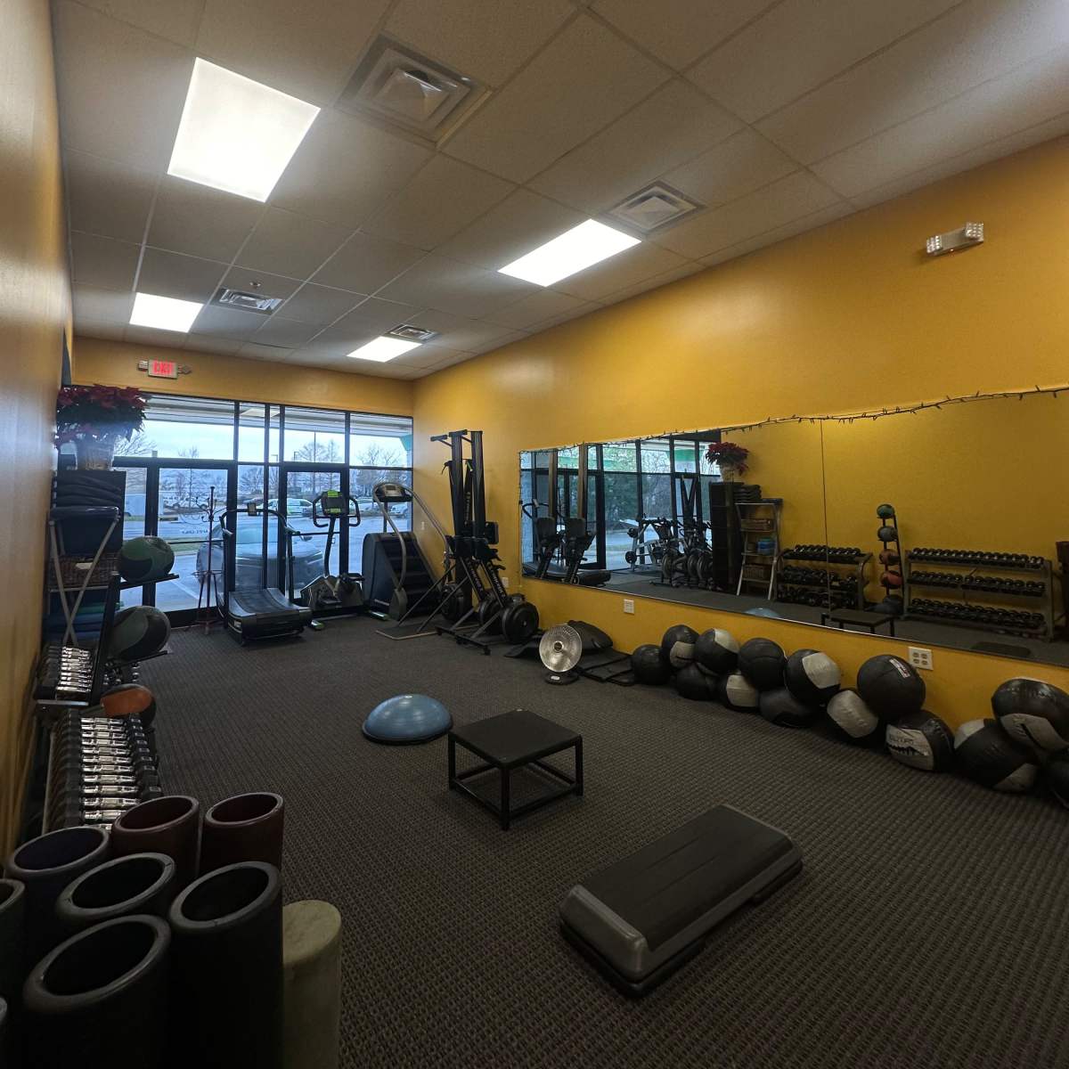 A fitness studio with exercise equipment, including weights and mats, alongside large windows allowing natural light.