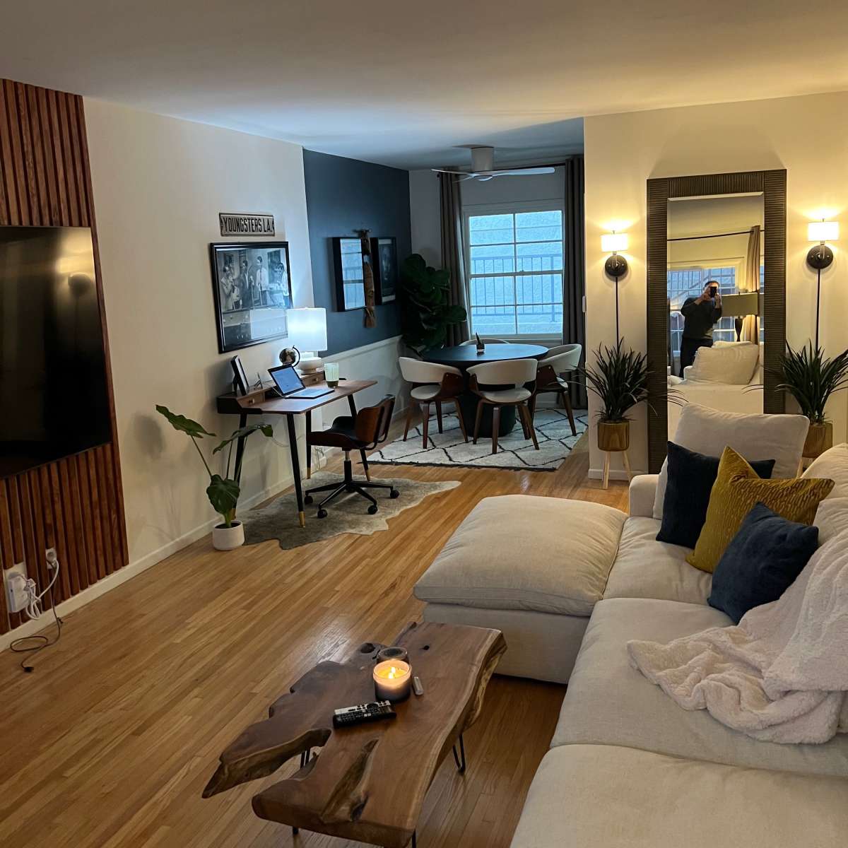 A modern living room features a beige sectional sofa, a wooden coffee table, dining area with a round table and chairs, and a workspace with a desk, all illuminated by natural light from large windows.
