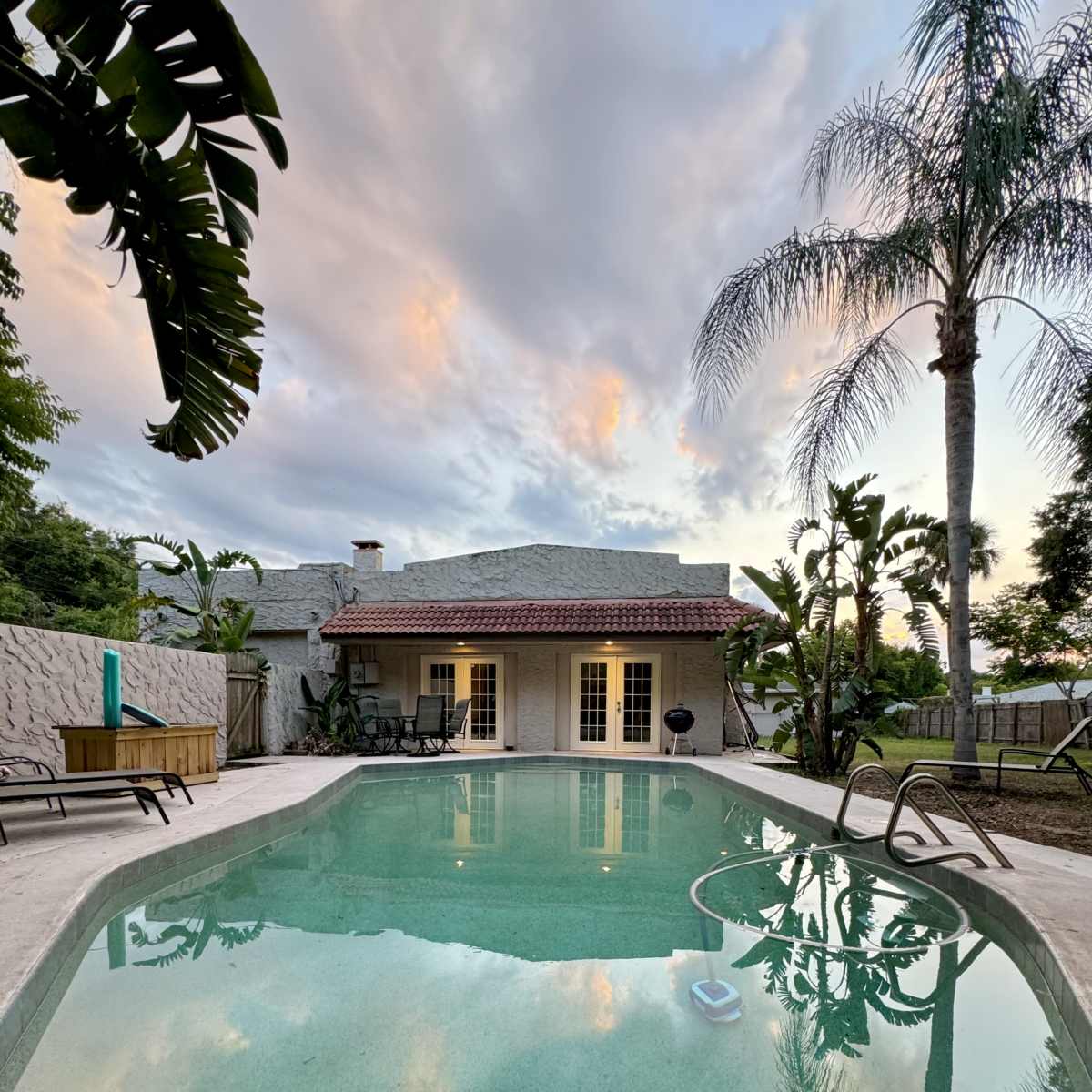A backyard scene features a swimming pool surrounded by a patio and tropical plants, with a house and cloudy sky in the background.