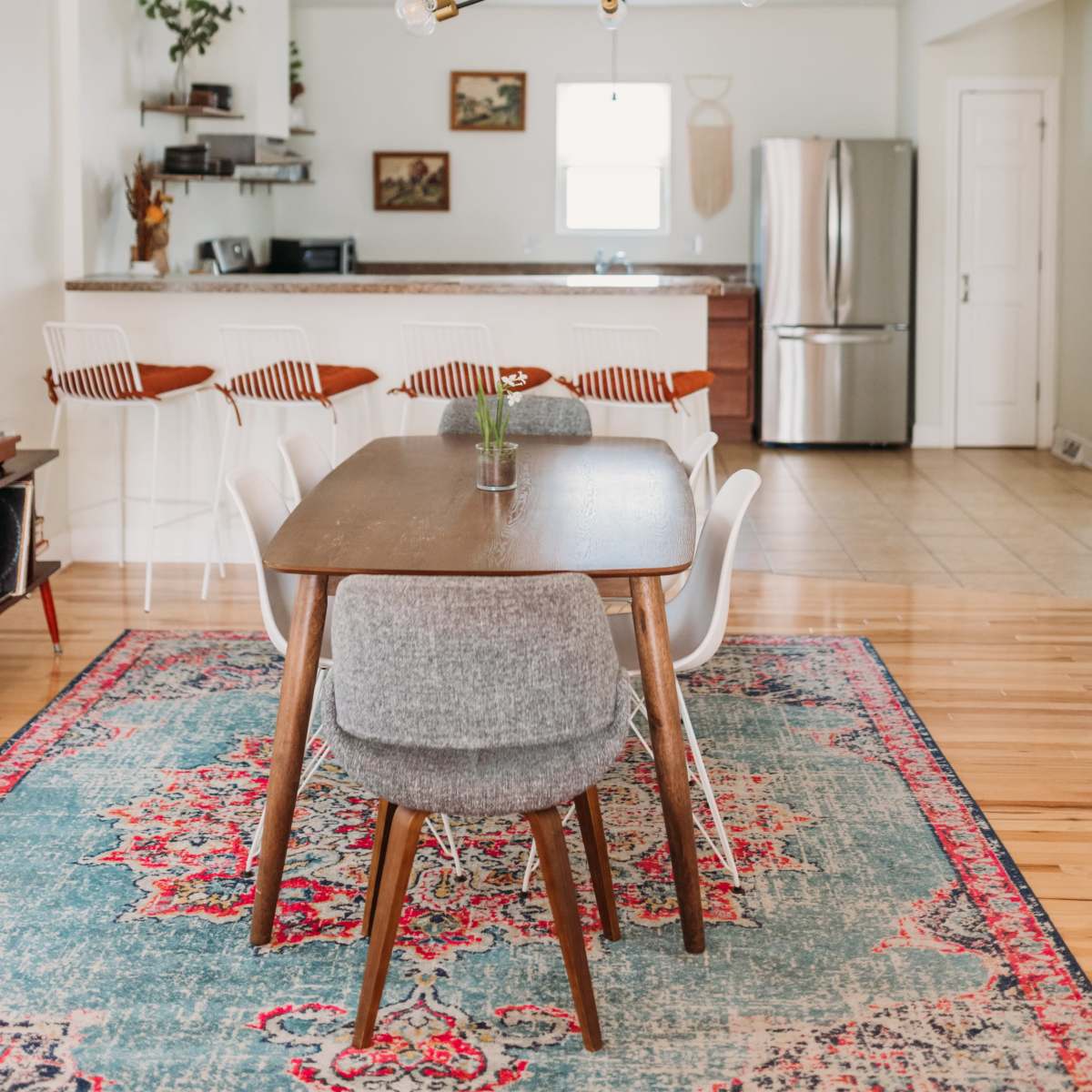 A dining area features a wooden table surrounded by chairs, a colorful rug, and a modern light fixture, with a kitchen in the background.