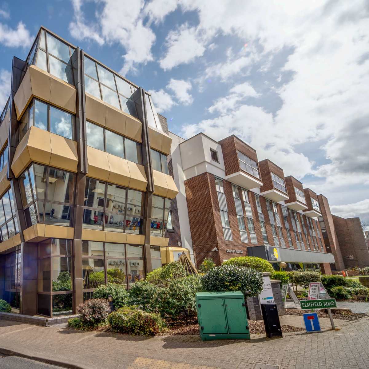 A modern, multi-level building with large glass windows and a gold exterior is situated next to a brick structure, surrounded by landscaped greenery under a partly cloudy sky.