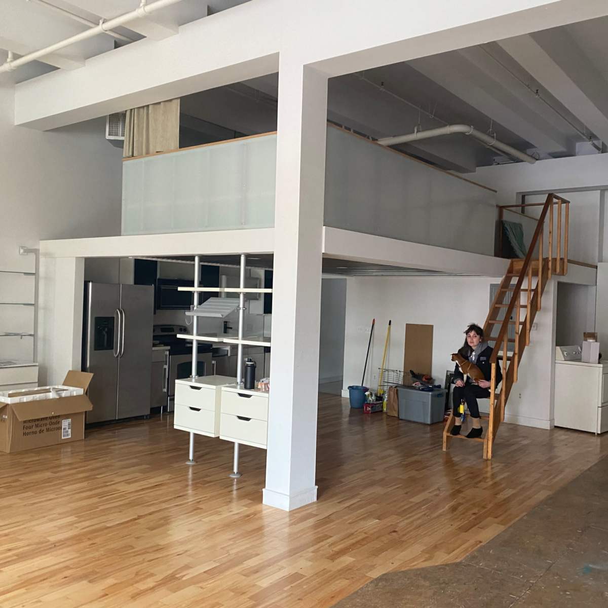 A person sits on a staircase in a spacious, partially furnished room with wooden floors and white walls.
