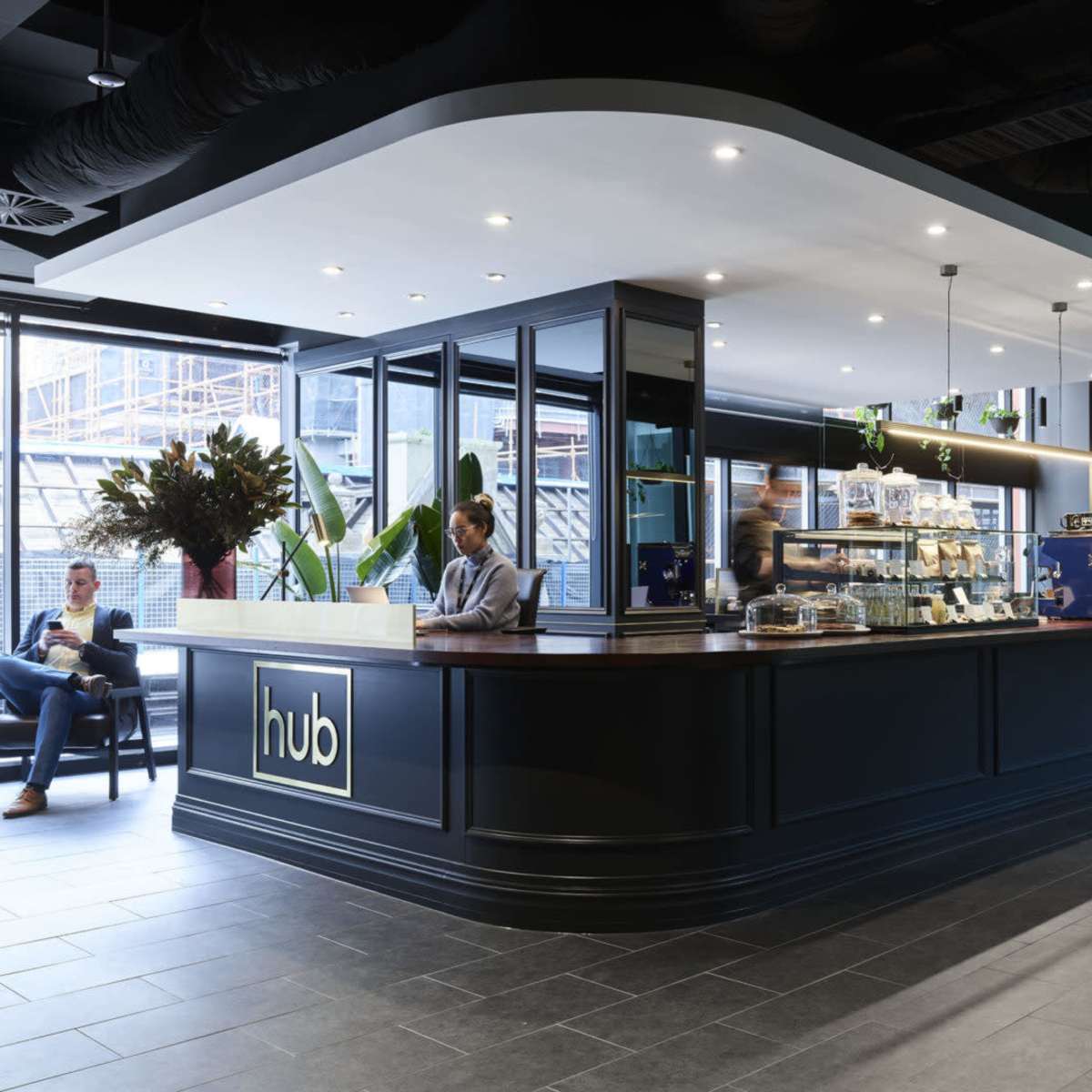 A modern café interior features a curved service counter, a barista preparing drinks, and seating areas surrounded by greenery.