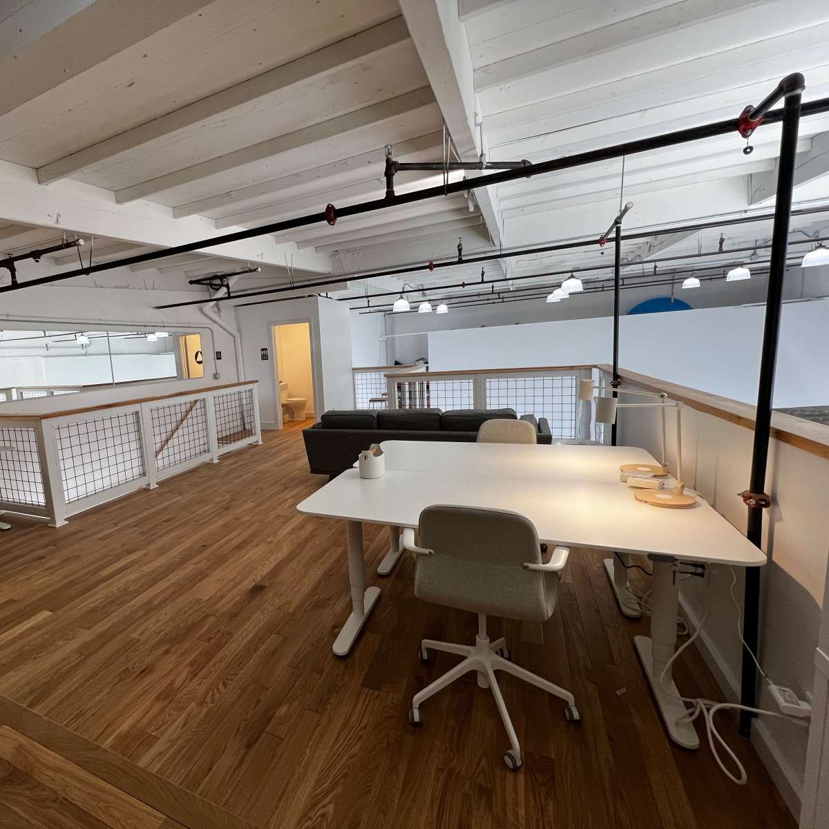 A spacious, modern office interior with wooden flooring, two workstations, a sofa, and overhead lighting.