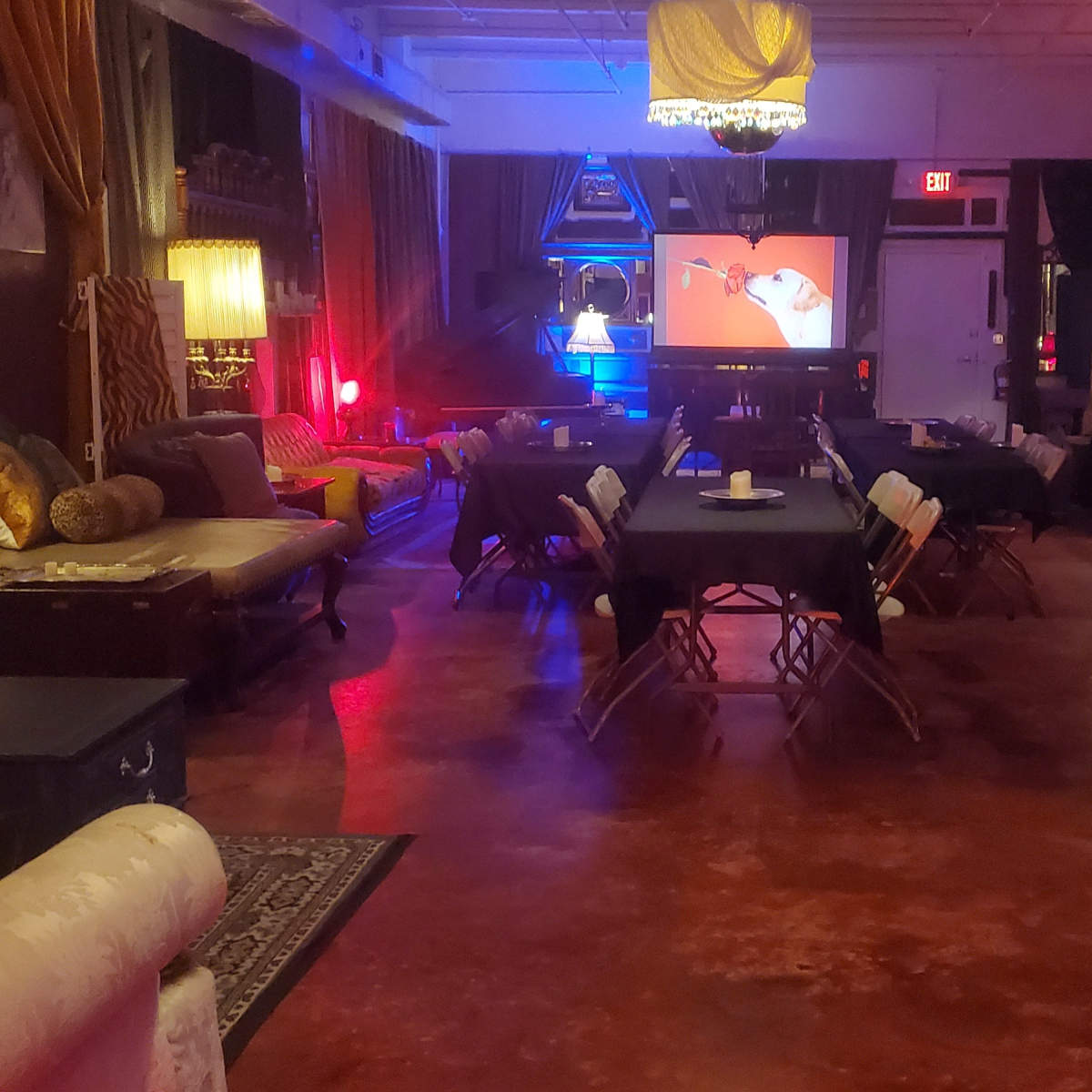 The scene shows a dimly lit event space with folding tables, chairs, and a large screen displaying a colorful image.