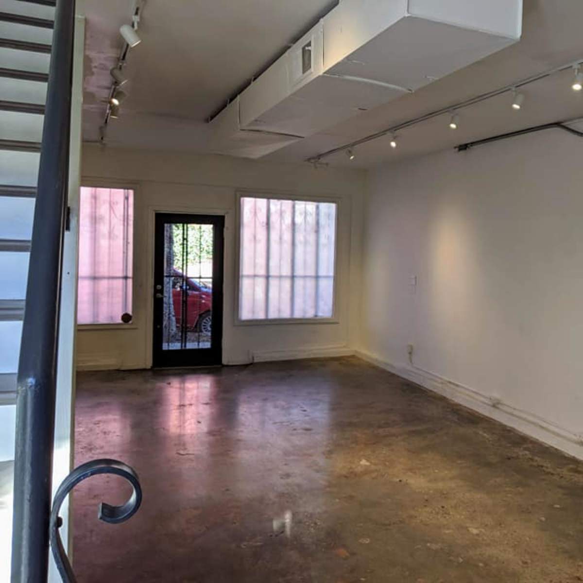 An empty gallery space with a concrete floor, white walls, and large windows on one side.
