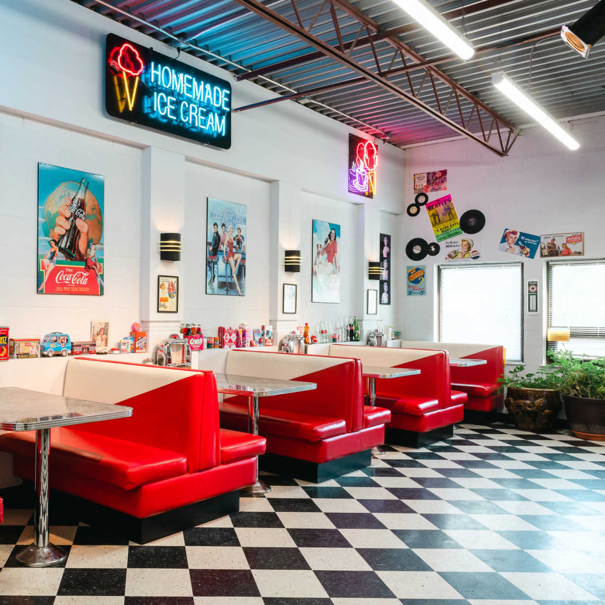 A retro-style diner features red upholstered booths and black-and-white checkered flooring, adorned with vintage advertisements and a neon sign for "Homemade Ice Cream."