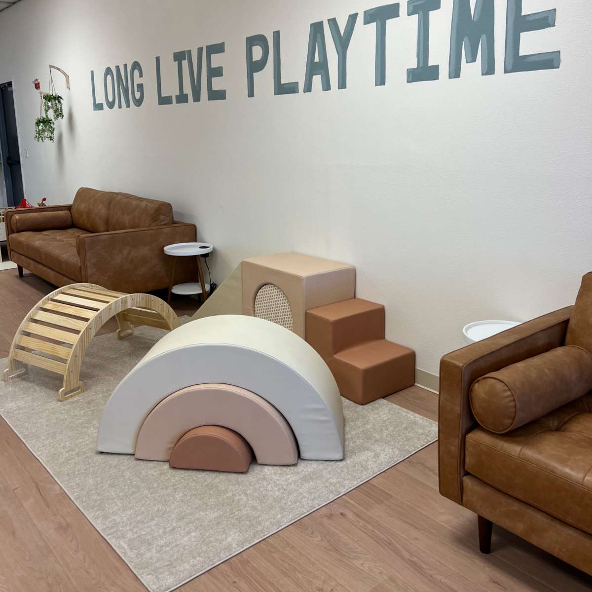 A play area with colorful soft play equipment, including a rainbow, ramps, and steps, alongside two brown couches and a wall that reads "LONG LIVE PLAYTIME."