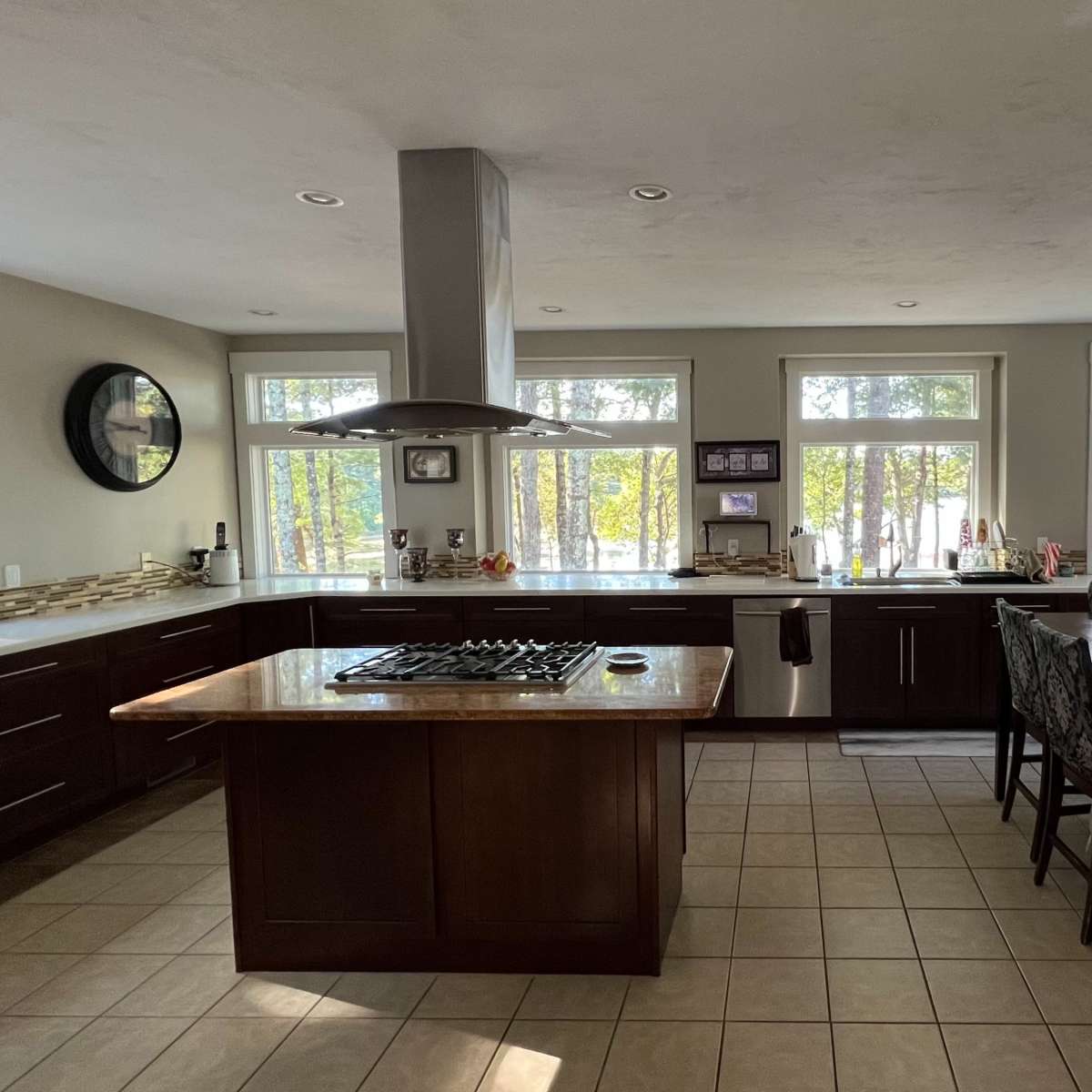 A modern kitchen features dark wood cabinets, an island with a gas stove, and large windows overlooking a wooded area.